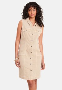 Abito in denim beige con colletto, chiusura frontale con bottoni e due tasche sul petto. Design senza maniche e lunghezza al ginocchio con cuciture visibili.