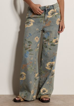 Ansicht von der Taille bis zu den Füßen einer Person, die weite Jeans mit gelbem und blauem Blumenmuster, braune Sandalen und ein weißes Oberteil trägt.