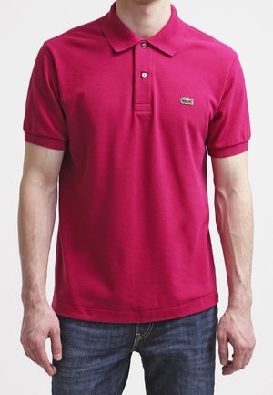 Mann trägt ein schlichtes dunkelrosa kurzärmeliges Poloshirt mit zwei Knöpfen und einem kleinen grünen Krokodillogo auf der Brust.