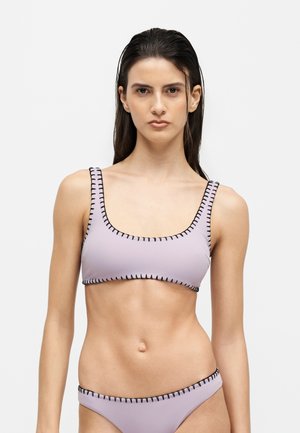 Junge Frau mit langen dunklen Haaren, die einen hellvioletten Bikini mit schwarzen, genähten Rändern trägt und vor einem einfarbigen hellen Hintergrund steht.