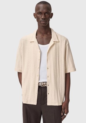 Mies, joka pukeutunut teksturoituun kerma-valkoiseen lyhythihaiseen nappipaitaan valkoisen topin päällä, tummiin housuihin, kuvioituun vyöhön ja hopeisiin sormuksiin oikeassa kädessä.