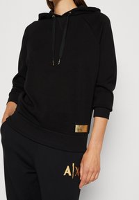 Felpa nera in morbido tessuto con zip in oro. Presenta un orlo e polsini a coste e una placca con logo dorato sul lato sinistro in basso.