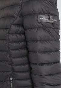 Giacca a vento nera con texture trapuntata e tasca con cerniera. Presenta una patch con il logo "FRIEDA FREDDIES" sulla spalla.