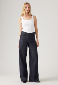 Σκούρο τζιν με φαρδιά γραμμή και ψηλή μέση, με πέντε τσέπες και μεταλλικά αξεσουάρ. Συνδυάζεται με ένα εφαρμοστό λευκό tank top.