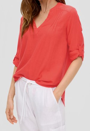 Koraalrode V-hals blouse met opgerolde mouwen en plooidetails. Gecombineerd met witte broek met trekkoord, met een ontspannen pasvorm en lichtgewicht stof.