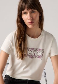 Cremefarbiges T-Shirt mit kurzen Ärmeln, das ein florales Muster innerhalb eines rosa Levi's-Logo-Graphics zeigt. Aus Baumwollmaterial, runder Rundhalsausschnitt, lässige Passform.