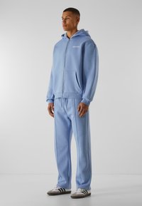Ljusblå zip-up hoodie med fickor och dragsko, tillsammans med matchande träningbyxor med sidofickor. Materialet är mjukt och slätt.