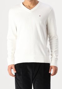 Muž oblečený v bílém svetr s výstřihem do V a dlouhými rukávy, s malým logem na hrudi, v kombinaci s černými kalhotami.