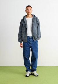 Felpa grigia con zip sopra una maglietta bianca a maniche lunghe, abbinata a jeans larghi blu e sneakers grigie. Design semplice, vestibilità comoda.