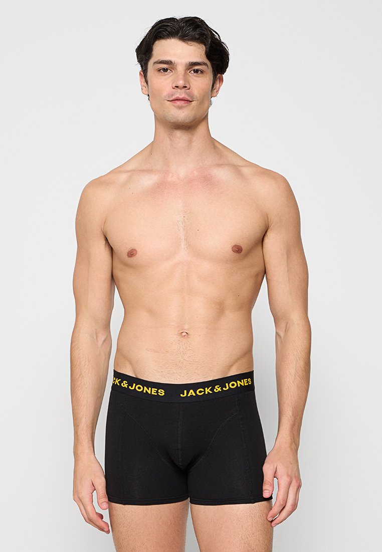 Hombre sin camisa que lleva bóxers negros de Jack & Jones con texto amarillo en la cinturilla, de pie frente a un fondo claro y liso.