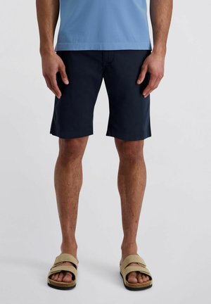 Mann in marineblauen Shorts, hellblauem Hemd und beigen Sandalen steht vor einem schlichten hellen Hintergrund.