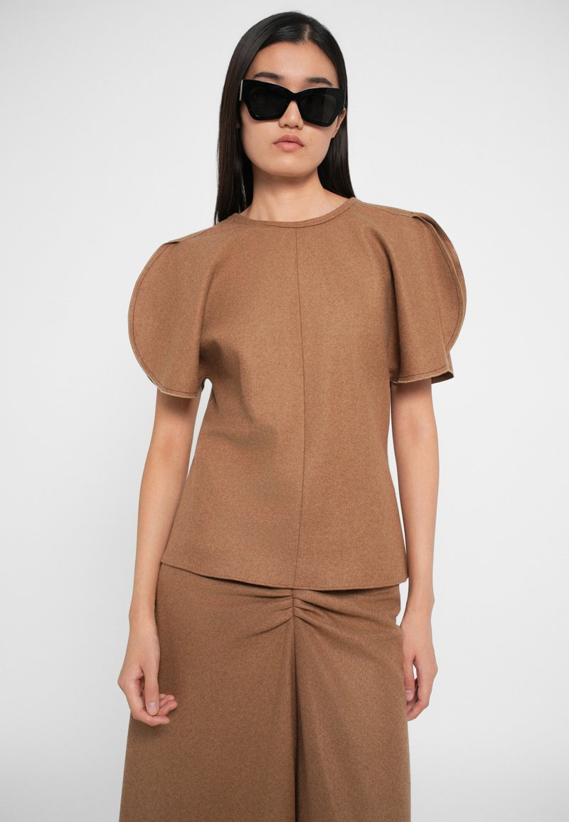 Top in maglina marrone con maniche a sbuffo, corsetto aderente e scollatura rotonda. Abbinato a una gonna raccolta coordinata. Modella che indossa occhiali da sole neri.