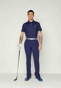 Ναυτική πολοχ shirt με μικρό λογότυπο, συνδυασμένο με ταιριαστά παντελόνια, γκρι ζώνη και μπλε αθλητικά παπούτσια; κρατώντας ένα μπαστούνι του γκολφ.