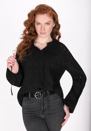 Blusa nera intrecciata con motivo a occhio di pernice, scollo arricciato e lacci. Maniche lunghe con polsini svasati. Abbinata a jeans neri aderenti e una cintura.