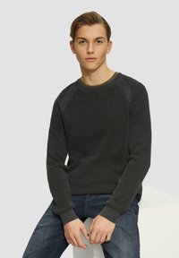 Dunkelgrauer strukturierter Pullover mit geripptem Kragen und Bündchen, getragen über einem grünen Hemd, kombiniert mit blauen Jeans, Modell sitzt auf einem weißen Sockel.