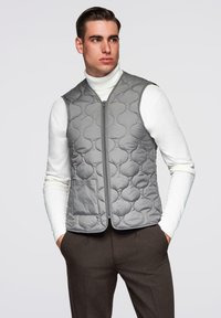 Mann trägt eine graue gesteppte Weste über einem weißen Rollkragenpullover und dunkelbraunen Hosen, steht mit den Händen in den Taschen.