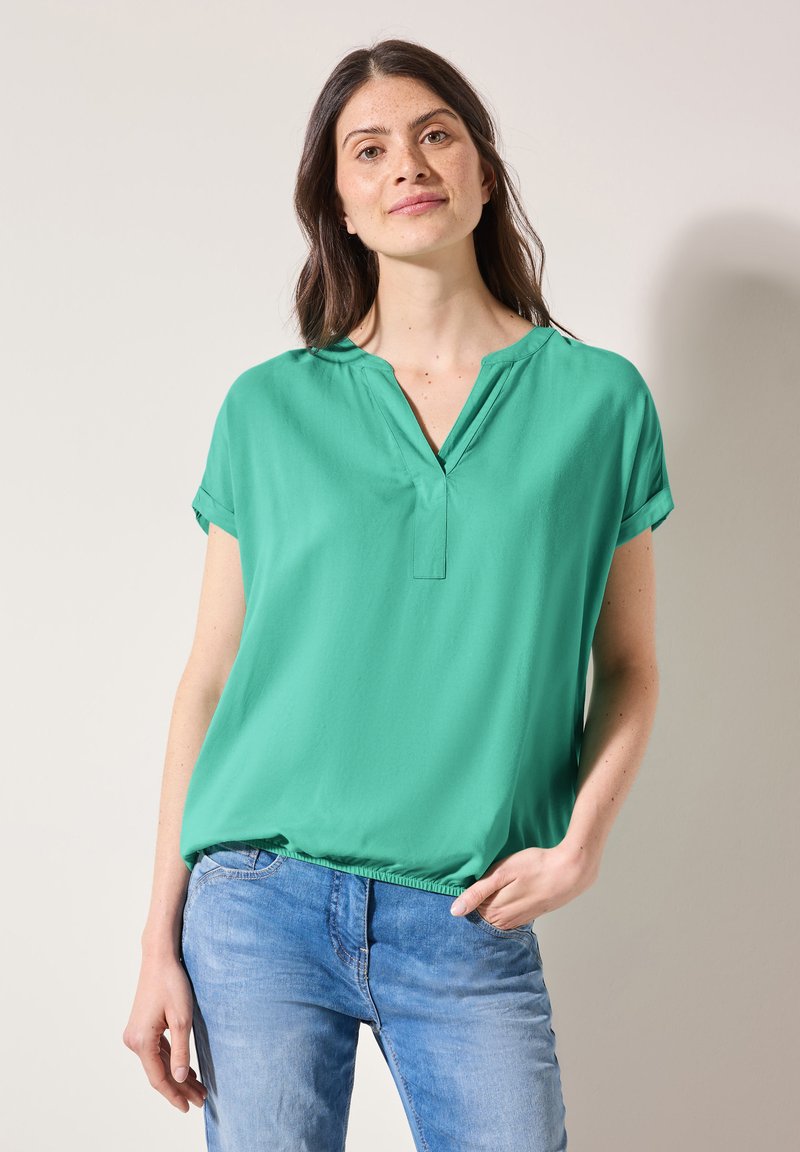 Grøn kortærmet bluse med V-hals og afslappet fit, lavet af glat stof. Båret med lyseblå jeans, hvilket giver et afslappet udseende.