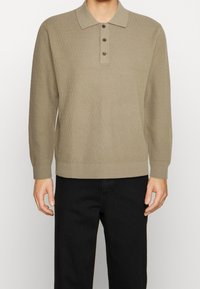 Polo lungo a maniche con lavorazione a maglia color beige, con un motivo testurizzato, tre bottoni marroni al collo e polsini a costine. Indossato con pantaloni neri.