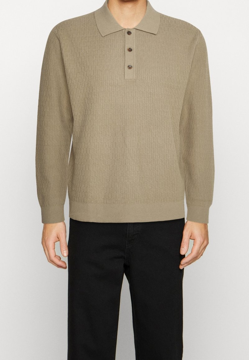 Polo lungo a maniche con lavorazione a maglia color beige, con un motivo testurizzato, tre bottoni marroni al collo e polsini a costine. Indossato con pantaloni neri.