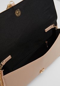 Beige laukku, joka on valmistettu keinonahasta, kultasävyisellä napilla ja ketjuhihnalla. Sisäpuoli on vuorattu mustalla kankaalla, ja siinä on vetoketjullinen tasku turvalliseen säilyttämiseen.