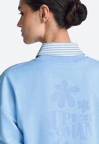 Hele sinine pusa, millel on kõrgendatud lillemuster ja tekst "SUPER WOMAN." Kraega sinise ja valge triibulise kangaga.