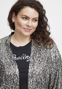 Fényes fekete és ezüst flitteres dzseki gallérral; egy fekete ing fölött viselve, amelyen ezüst írásban a "Feeling" felirat található. Közepes hosszúságú göndör haj.