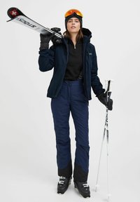 Slidinėjimo įranga, apimanti tamsią striukę, juodą vidurinį sluoksnį, tamsiai mėlynas kelnes, juodus slidinėjimo batus, slidinėjimo lazdas ir apsauginius akinius su oranžiniu tonu.