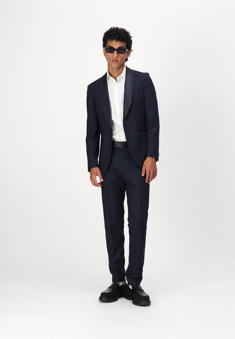 Costume marine avec un revers en satin, chemise blanche en dessous, accompagné de lunettes de soleil noires et de chaussures noires. Un pantalon ajusté complète le look.