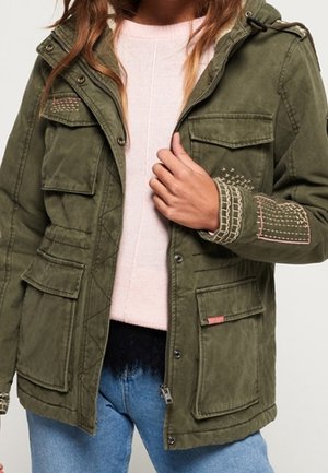 Woman wearing an olive green jacket with embroidered patches on sleeves, multiple flap pockets, over a light pink sweater and blue jeans.