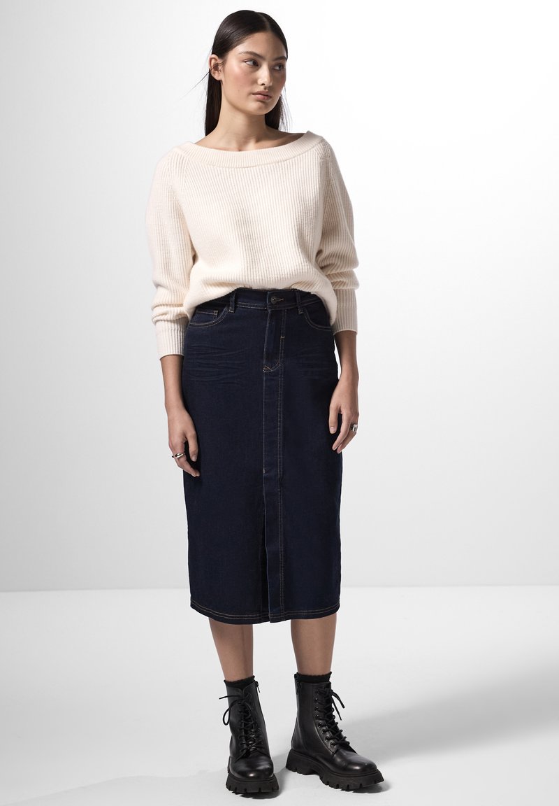 Suéter de punto acanalado beige con escote amplio, combinado con una falda midi de mezclilla oscura que presenta un cierre frontal y costuras en contraste. Botas negras de cordones.