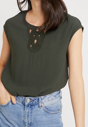 Dunkelgrünes ärmelloses Oberteil mit filigranem Cut-out- und Stickdesign am Ausschnitt, kombiniert mit hellblauen Jeans.