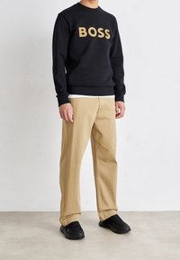 Hombre vistiendo un sudadera negra con "BOSS" en texto beige, pantalones beige y zapatillas negras, de pie contra una pared blanca.