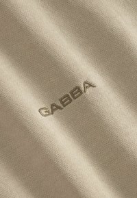 Beige strikket stof med det broderede logo "GABBA" i en lidt mørkere beige, der viser en glat og blød tekstur.