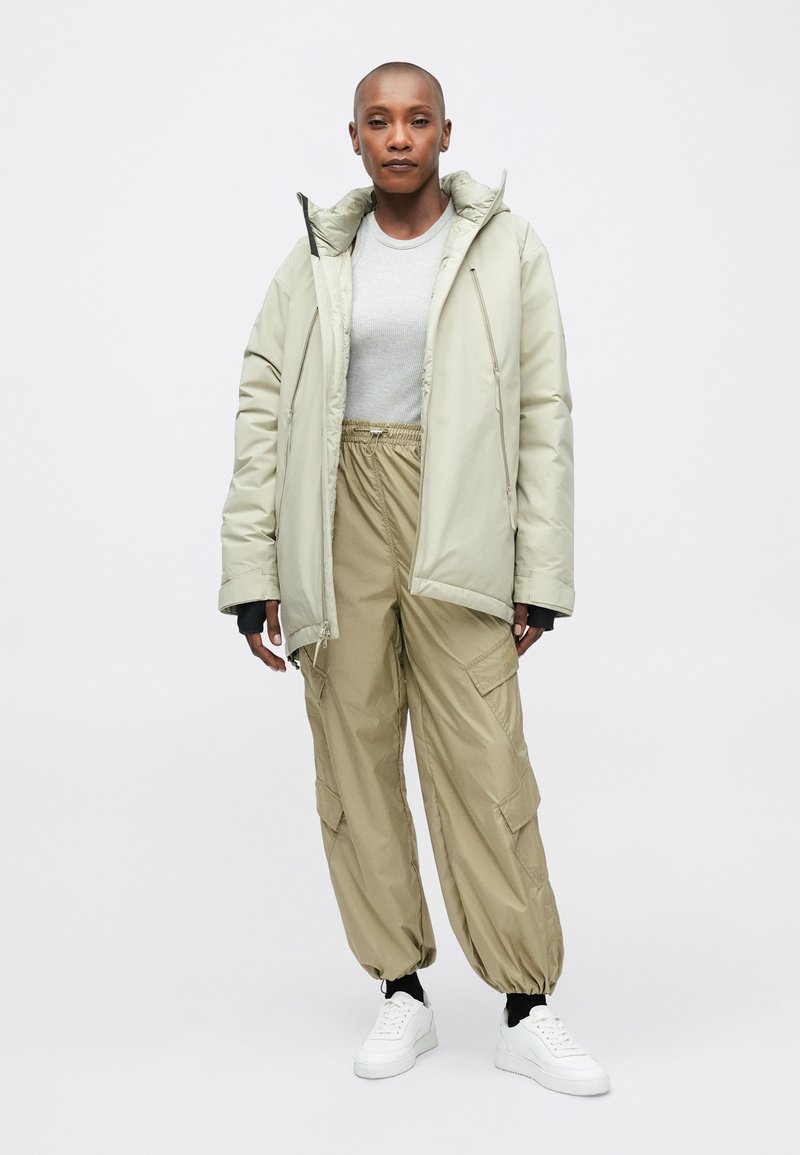 Beige Pufferjacke mit Kapuze, kombiniert mit lockeren Cargo-Hosen. Weiße Sneakers. Weicher Stoff, einfaches Design und neutrale Farbpalette.