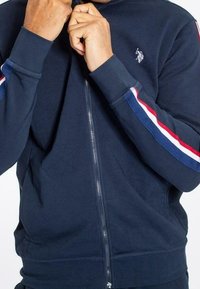 Hombre vestido con una chaqueta de cremallera azul marino con mangas a rayas en rojo, blanco y azul, y un pequeño logotipo bordado en blanco en el pecho.