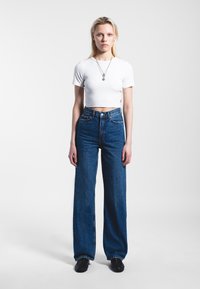 White cropped t-shirt and high-waisted, wide-leg denim jeans in dark blue. Simple silver necklace. Black shoes. Plain white background.