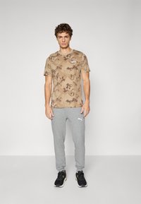 T-shirt à motif camouflage beige avec un petit logo, associé à un pantalon de jogging gris portant un logo de marque, et des chaussures de sport noires.