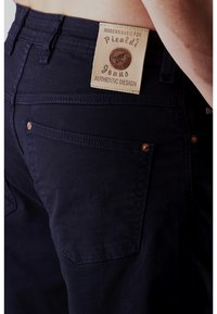 Dark blue jeans with a leather label featuring "Authentic Design." Notable accents include copper hardware and contrast stitching.
