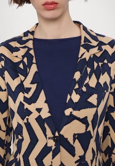 Beige blazer with navy zigzag pattern, textured fabric, notched collar, single button closure, layered over a navy top.