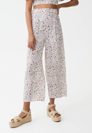 Witte wijde broek met een paars en zwart gespikkeld patroon, hoge taille en een knoopsluiting; gecombineerd met lichtbeige wedge sandalen.
