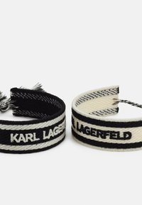 Mustvalge kudumistega käevõrud, millel on kontrastsetes värvides tikitud "KARL LAGERFELD", ja millel on selgelt eristuvad triibud ning kulunud servad.