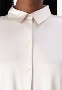 Kreemikas särgitüüp blouse, millel on satiinist krae, esipesul ja sile tekstuur, loodud minimalistlikku esteetikat silmas pidades.