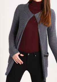 Žena v šedém kardiganu s texturou, který má otevřený přední díl, přes burgundské rolákové svetry a černé kalhoty.