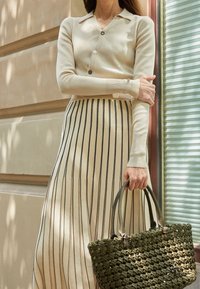 Cárdigan color beige con cuello y cierre de botones, combinado con una falda beige con rayas verticales negras, sosteniendo un bolso de mano de mimbre verde.