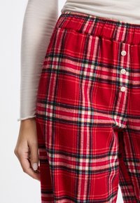 Czerwone dresowe spodnie z flaneli w wzór tartan z białymi i czarnymi liniami. Elastyczny pas z guzikami do regulacji. Miękka tekstura.