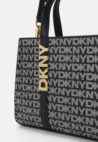 Czarna i biała torebka z wzorem logo DKNY, z elementami metalowymi w kolorze złotym, teksturowanym materiałem oraz sztywną konstrukcją i paskami na ramię.