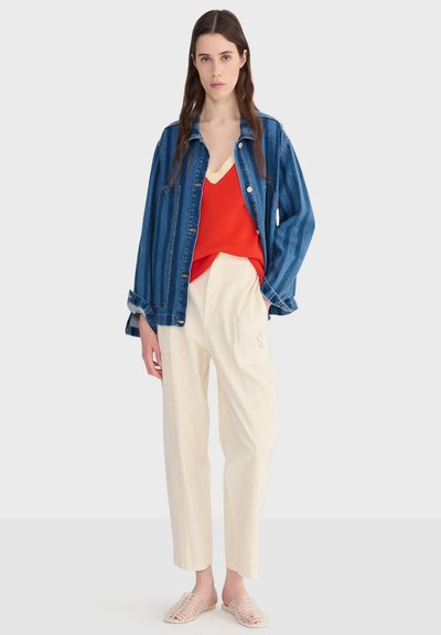 Femme aux longs cheveux bruns portant une veste en jean bleue, un pull rouge à col en V, un pantalon crème et des chaussures beige tissées sans lacets, debout sur un fond blanc.