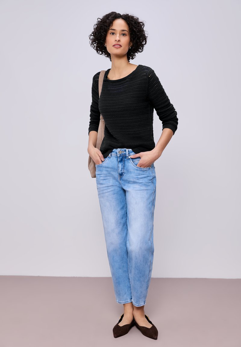 Femme aux cheveux bouclés portant un pull en maille noir, un jean bleu clair, des ballerines pointues noires et un sac à épaule beige, debout les mains dans les poches.