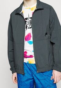 Grijze lichtgewicht jas met een rits aan de voorkant en elastische manchetten, gedragen over een kleurrijk tie-dye shirt en blauw met patroon shorts. Nike-logo zichtbaar.