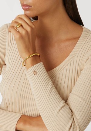 Gold-toned metal jewelry featuring a wavy cuff bracelet and a twisted ring, worn with a long-sleeve ribbed beige top.